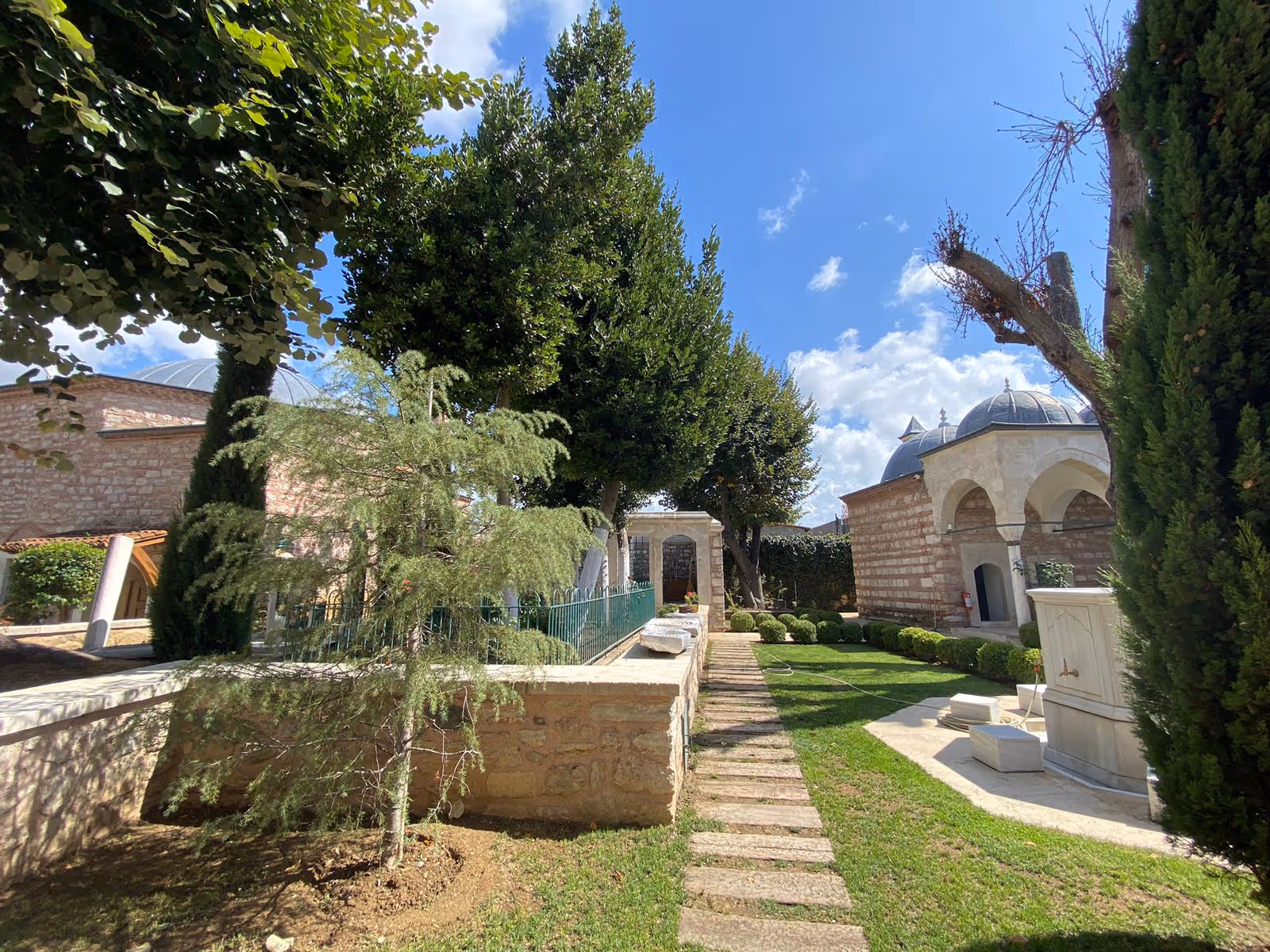 Sunlit garden with trimmed bushes, trees, stone pathways, and historic stone buildings with domed roofs under a blue sky with clouds.